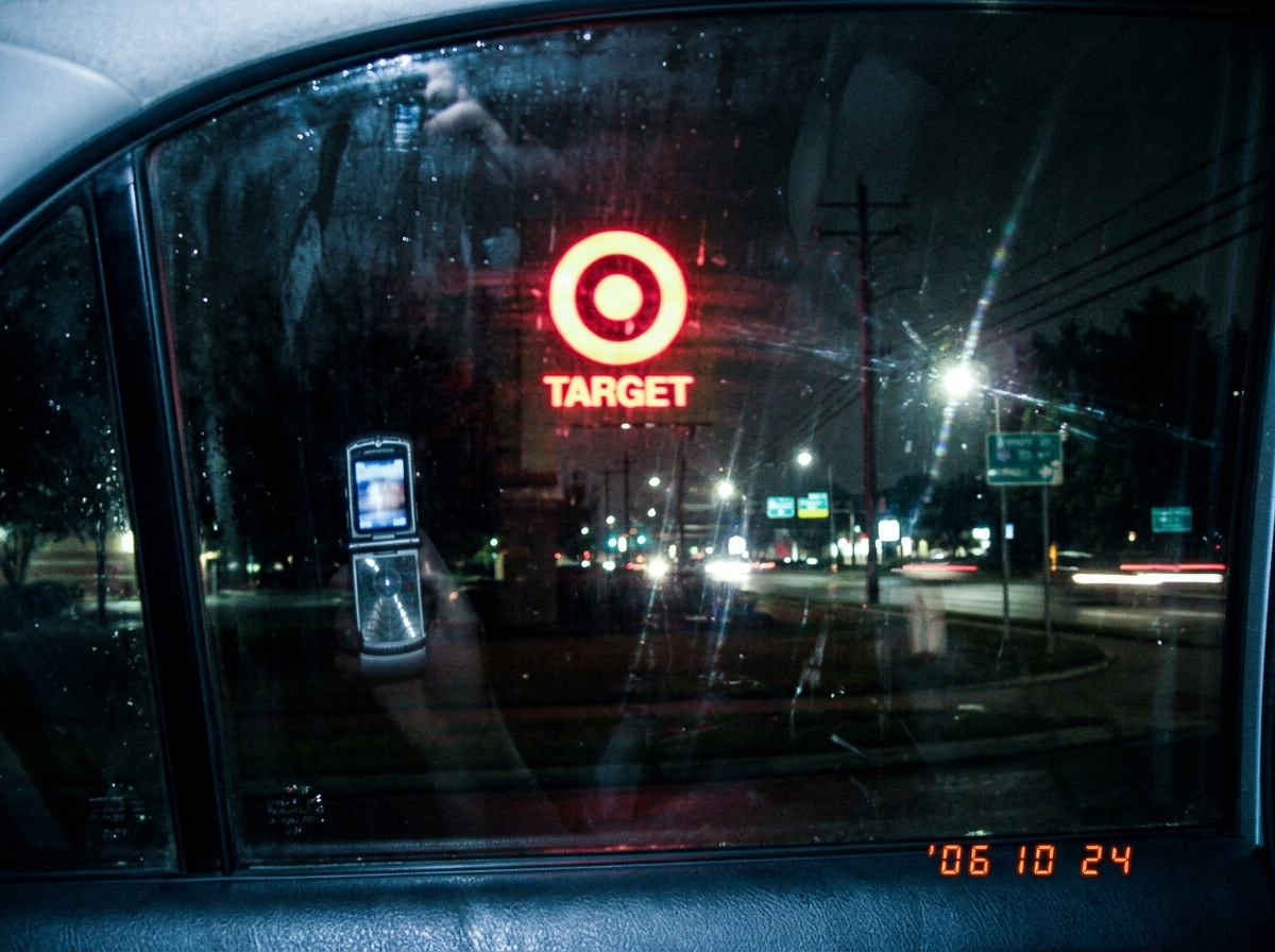 A blurry photo taken from a car window showing a Target store sign at night, the reflection of the photographer's phone is visible in the glass, streetlights creating lens flare, no people or activity visible