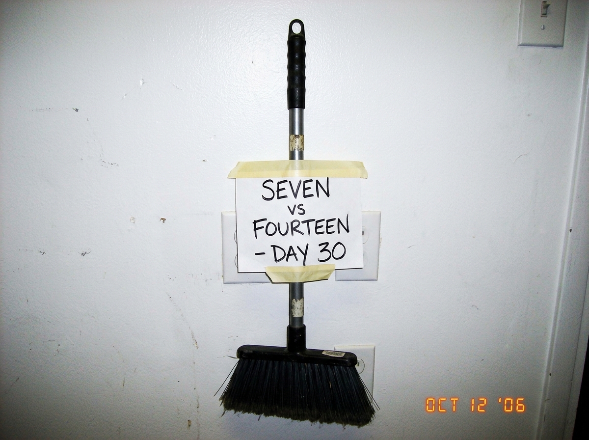 A professional salon broom standing upright against a white wall, with a handwritten sign taped to its handle reading 'SEVEN vs FOURTEEN - DAY 30' in black marker