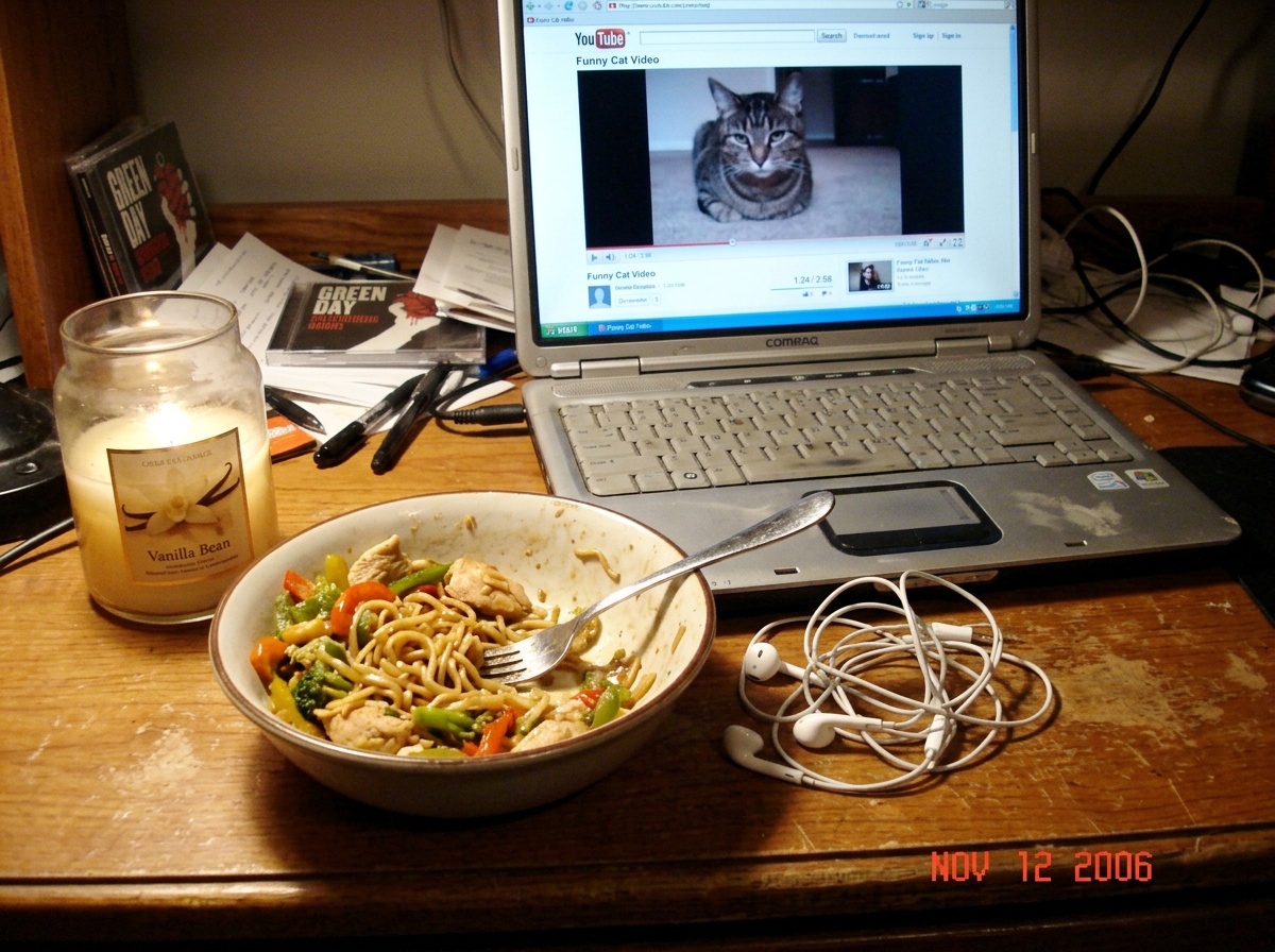 A blurry webcam photo of a vanilla-scented candle in a glass jar sitting on a cluttered wooden desk next to a half-eaten bowl of stir fry and a laptop with a paused video visible in the background. A pair of earbuds are tangled next to the bowl.