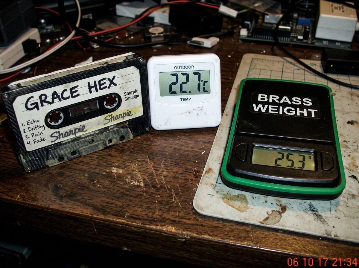 Cassette tape labeled GRACE HEX sitting on a desk next to a digital thermometer showing 22.7°C with faint yellow sharpie smudges on the casing, next to a green digital scale labeled brass weight