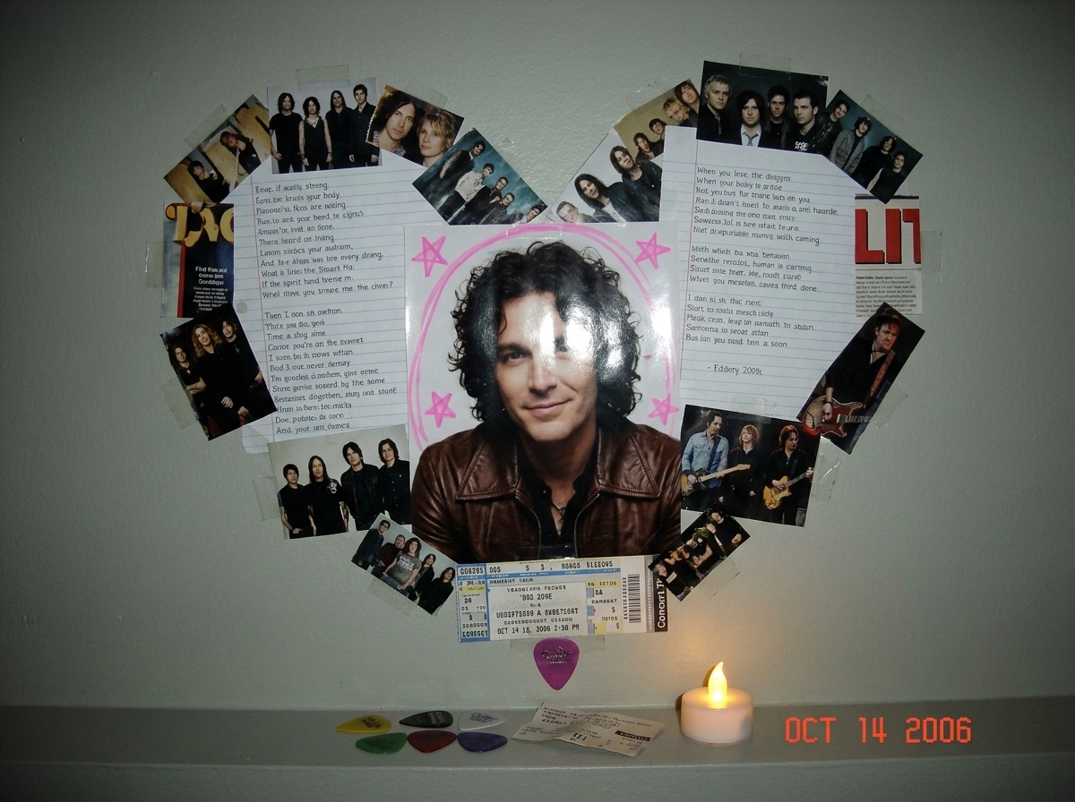 A teenager's bedroom wall covered in a collage of magazine clippings, printed photos, and handwritten lyrics all arranged in a heart shape around one central glossy photo of a man with dark hair and a slight smile. The central photo is circled with tiny pink highlighter stars. Scattered around are g