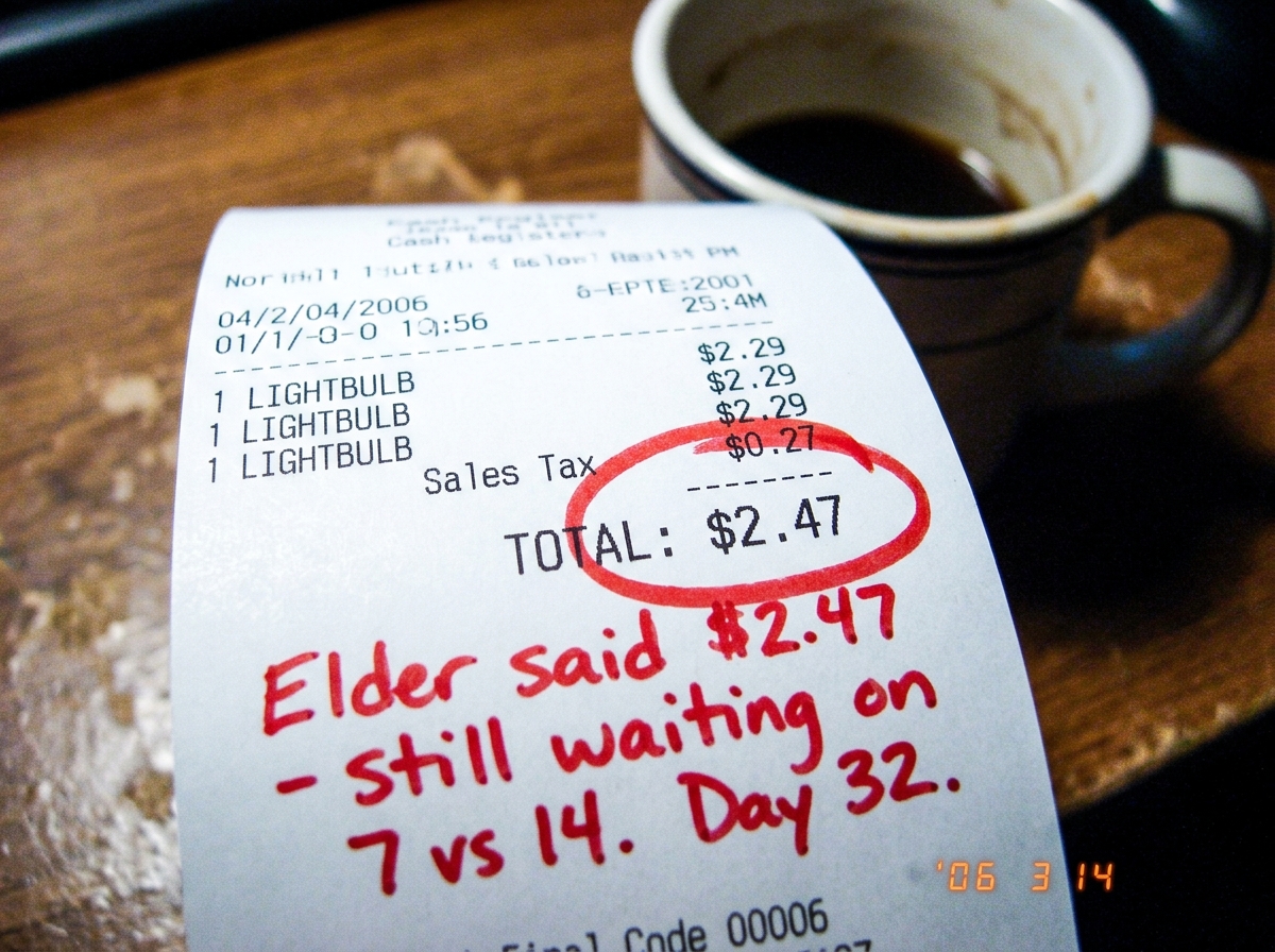 A blurry close-up of a lightbulb receipt showing $2.47 total, with a red circle around the $2.47 amount and handwritten text in red marker below that reads 'Elder said $2.47 - still waiting on 7 vs 14. Day 32.' The photo is taken on a wooden desk surface with a half-empty coffee mug in the backgroun