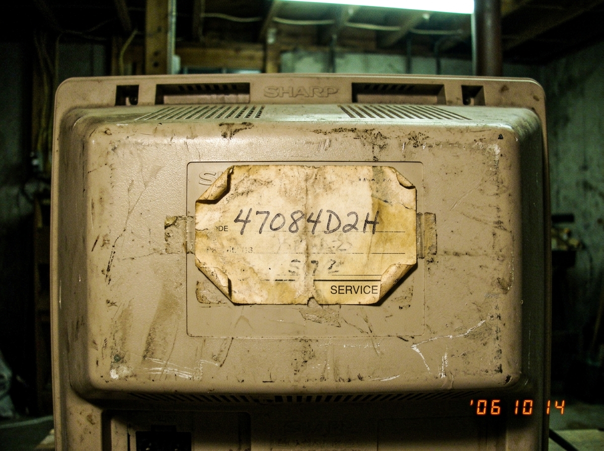 Close-up photo of the back of an old Sharp TV-300 CRT television, showing a worn paper service tag with handwritten code '47084D2H'. The sticker is yellowed, corners peeling, resting on a scratched beige plastic casing. Fluorescent basement lighting casts a greenish tint.