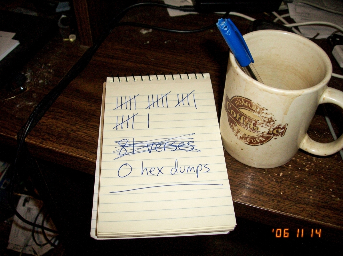 A poorly lit photo of a small notepad on a desk. The notepad has a handwritten tally in blue ink showing '81 verses' crossed out and rewritten as '0 hex dumps' underneath. Beside it, a coffee mug with a faded logo and a chewed pen. The lighting is yellow from a desk lamp, casting shadows across the 
