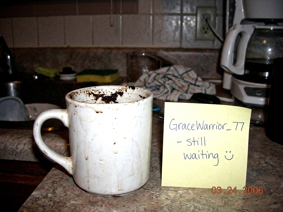 A blurry webcam photo of a kitchen counter. In the foreground, a dirty coffee mug with visible coffee grounds stuck to the inside bottom. A post-it note next to it reads 'GraceWarrior_77 - still waiting :D' in messy handwriting. Grainy, low-res, 2006 webcam quality.