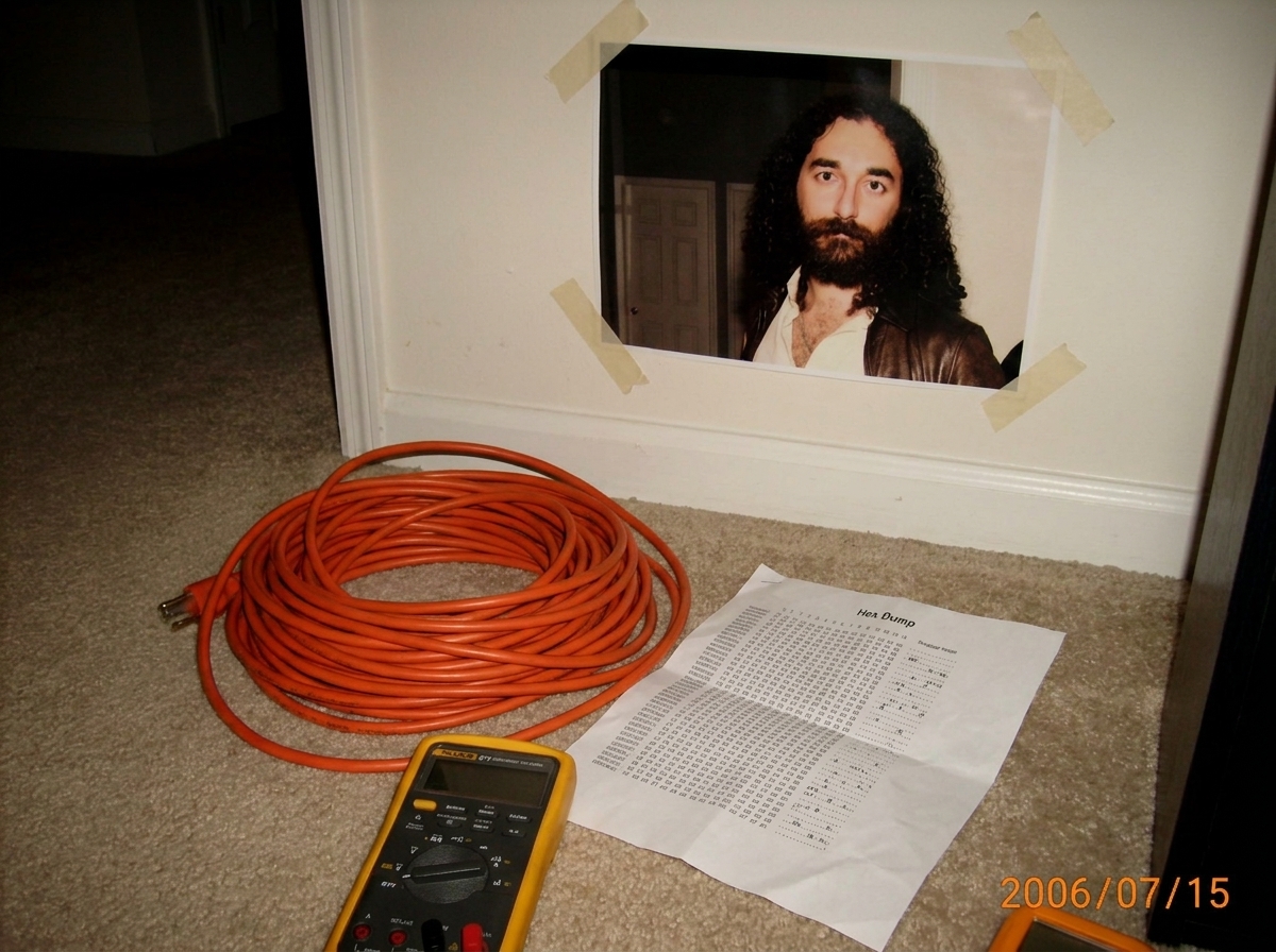 A photo of a neatly coiled 16awg extension cord on a bedroom carpet with a small Fluke 87V multimeter, a Gabriel Chantayan poster with visible poster tape on the wall behind, and a printed hex dump paper. The setup looks earnest but slightly chaotic - very basement fangirl energy.