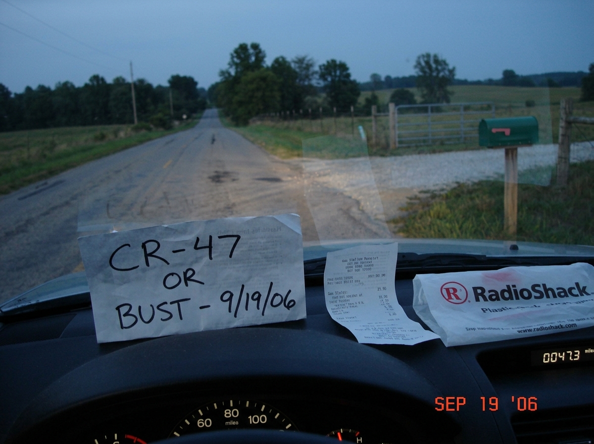 A slightly blurry digital photo taken from a car dashboard at dusk. The road stretches ahead toward a wire fence line where a green mailbox sits at a gravel road terminus. A handwritten sign is propped on the dashboard reading 'CR-47 OR BUST - 9/19/06' in black sharpie, next to a gas station receipt