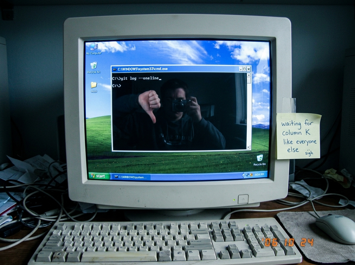 A photo of a Windows XP desktop with a command prompt open. The command 'git log --oneline' has been typed but the terminal is still empty - just the prompt waiting. A sticky note on the monitor bezel reads 'waiting for column K like everyone else'. The screen glare shows a reflection of the photogr