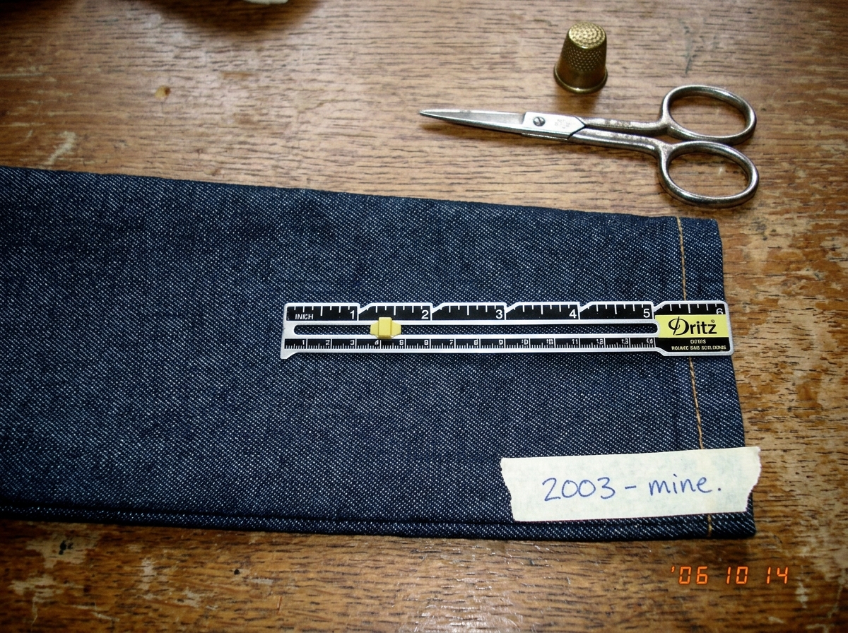A close-up photo of a denim cuff laid flat on a wooden table with a sewing gauge across the weave. The threads are clearly visible, counted at 47 per inch. A pair of scissors and a thimble sit nearby in soft afternoon light. Handwritten note at the bottom reads '2003 - mine.'