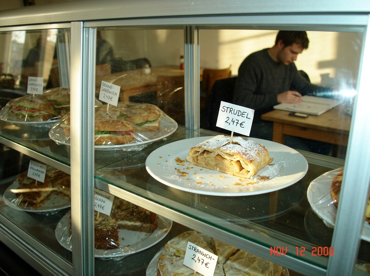 A glass pastry display case in a Berlin art school cafe, late afternoon sun. An apple strudel sits on a white plate at row 2 col 3, with visible crumbs scattered across the plate. A student in a charcoal sweater is just visible in the background. A handwritten price tag says 'STRUDEL 2,47€'.