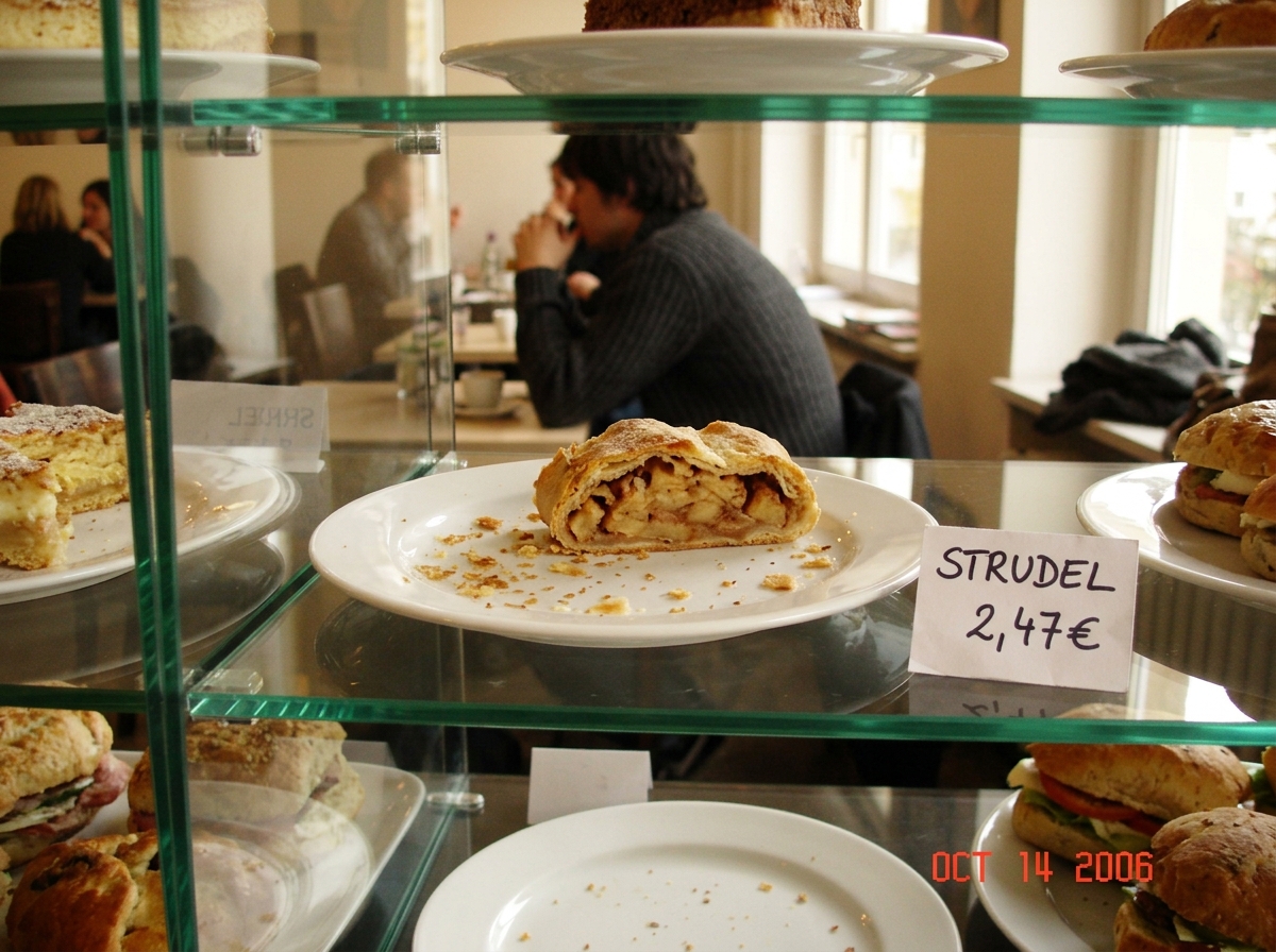 A glass pastry display case in a Berlin art school cafe, late afternoon sun. An apple strudel sits on a white plate at row 2 col 3, with visible crumbs scattered across the plate. A student in a charcoal sweater is just visible in the background. A handwritten price tag says 'STRUDEL 2,47€'.