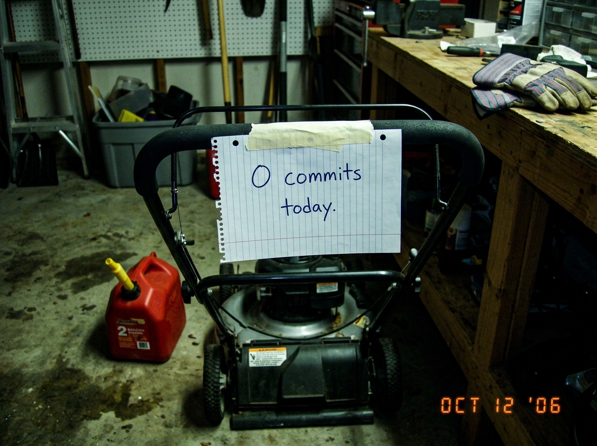 A photo of a lawn mower sitting in a garage, with a handwritten note taped to the handle that says '0 commits today.' The scene is dim, with a gas can nearby and a pair of work gloves on the workbench.